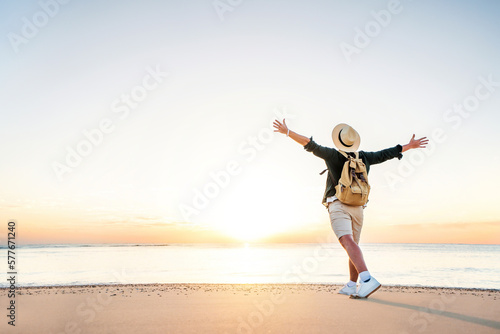 Happy man wearing hat and backpack raising arms up on the beach at sunset - Delightful man enjoying peaceful moment walking outdoors - Wellness, healthcare, traveling and mental health concept
