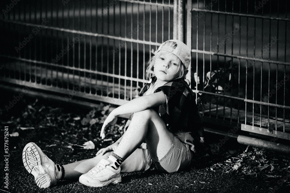 Obraz premium Black and white portrait of a stylish blonde model child in casual clothes and a cap sits on the ground leaning against a fence