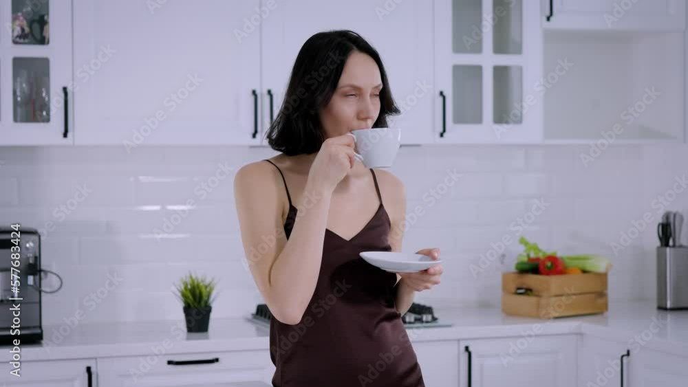 Middle-aged brunette woman in a dress drinking coffee or tea in the kitchen Middle-aged brunette woman in a dress drinking coffee or tea in the kitchen