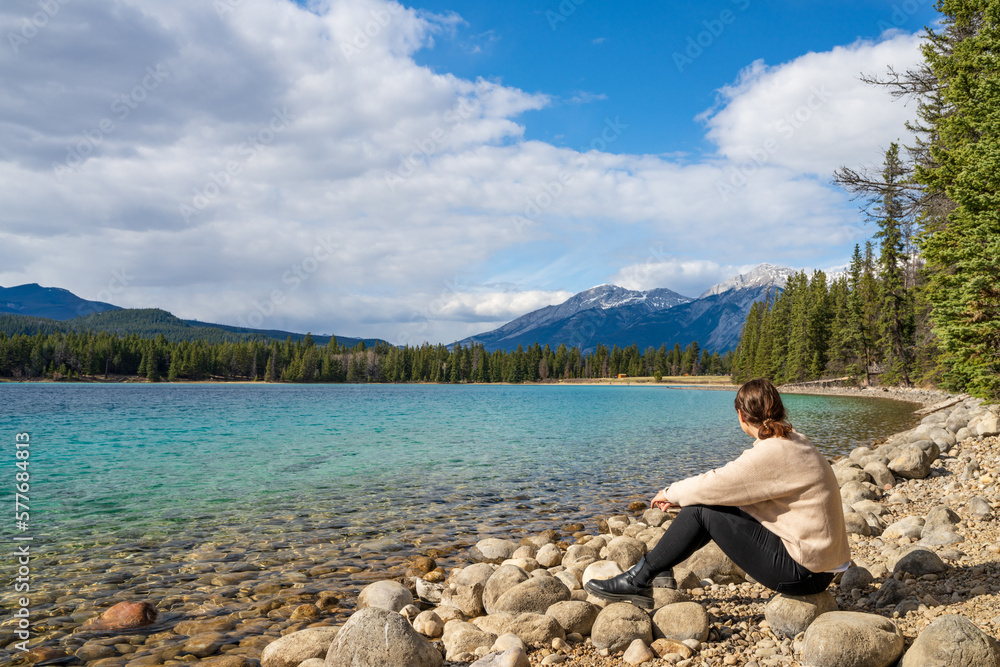 Young girl sitting by the lake shore enjoying the nature scenery. Summer time in Lake Annette, Jasper National Park, Canadian Rockies, Alberta, Canada.