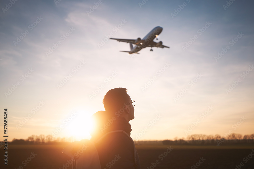 Man with backpack looking up to airplane landing at airport during beautiful sunset. .. - obrazy, fototapety, plakaty 