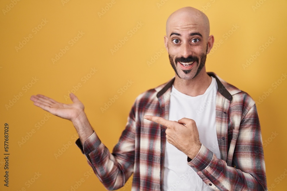 Hispanic man with beard standing over yellow background amazed and smiling to the camera while presenting with hand and pointing with finger. Hispanic man with beard standing over yellow background amazed and smiling to the camera while presenting with hand and pointing with finger.