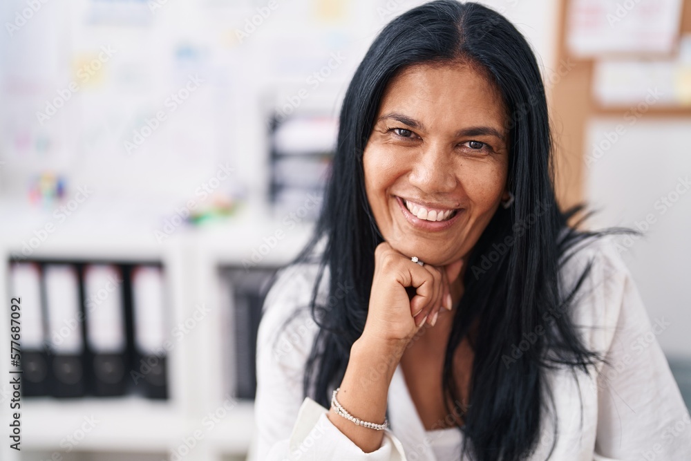 Middle age hispanic woman business worker smiling confident sitting on table at office