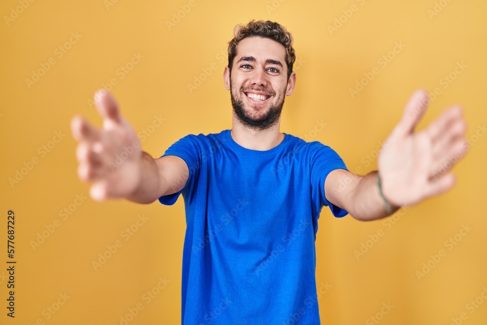 Hispanic man with beard standing over yellow background looking at the camera smiling with open arms for hug. cheerful expression embracing happiness. Hispanic man with beard standing over yellow background looking at the camera smiling with open arms for hug. cheerful expression embracing happiness.