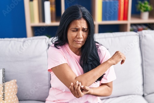Middle age hispanic woman sitting on sofa suffering for elbow pain at home