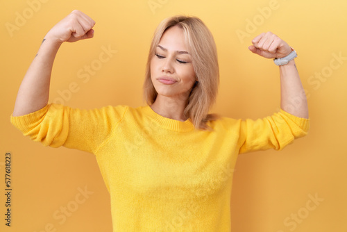 Young caucasian woman wearing yellow sweater showing arms muscles smiling proud. fitness concept.