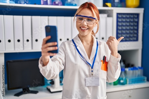 Young caucasian woman working at scientist laboratory doing video call with smartphone pointing thumb up to the side smiling happy with open mouth