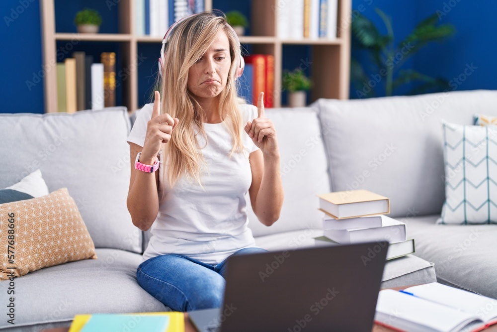 Young blonde woman studying using computer laptop at home pointing up looking sad and upset, indicating direction with fingers, unhappy and depressed. Young blonde woman studying using computer laptop at home pointing up looking sad and upset, indicating direction with fingers, unhappy and depressed.
