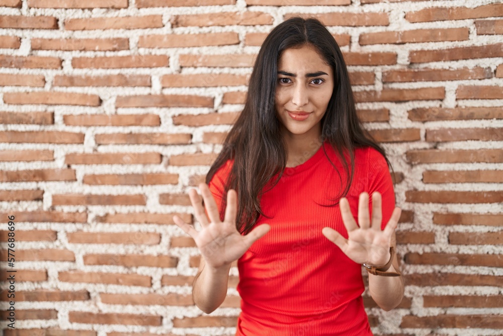 Young teenager girl standing over bricks wall moving away hands palms showing refusal and denial with afraid and disgusting expression. stop and forbidden. Young teenager girl standing over bricks wall moving away hands palms showing refusal and denial with afraid and disgusting expression. stop and forbidden.