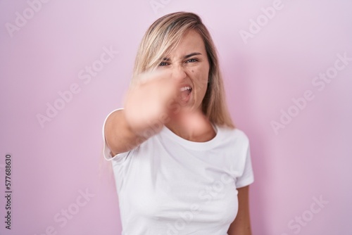 Young blonde woman standing over pink background pointing displeased and frustrated to the camera, angry and furious with you