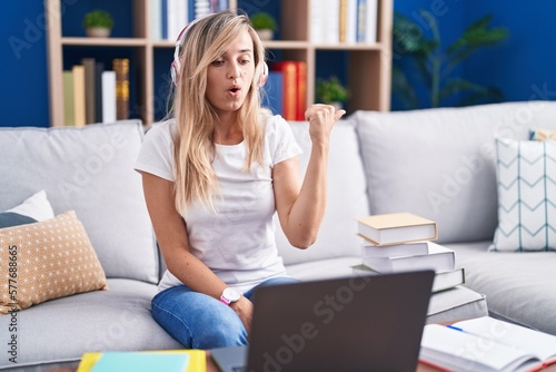 Young blonde woman studying using computer laptop at home surprised pointing with hand finger to the side, open mouth amazed expression.