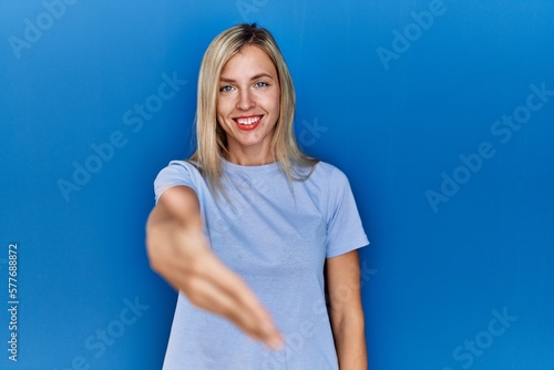 Beautiful blonde woman wearing casual t shirt over blue background smiling friendly offering handshake as greeting and welcoming. successful business.