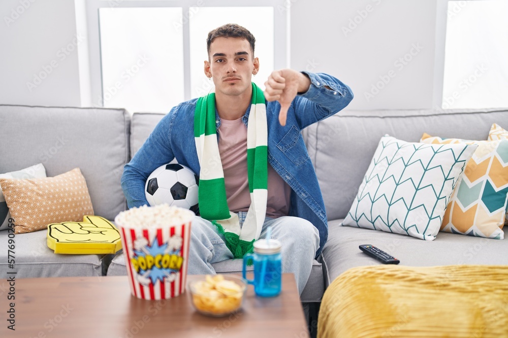 Handsome hispanic man holding ball supporting team with angry face, negative sign showing dislike with thumbs down, rejection concept Handsome hispanic man holding ball supporting team with angry face, negative sign showing dislike with thumbs down, rejection concept
