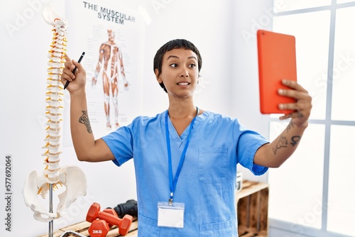 Young hispanic woman wearing physiotherapist uniform having video call pointing to vertebral column at physiotherapy clinic photo