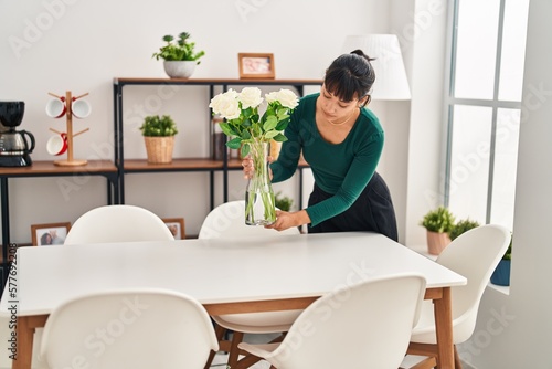 Young beautiful hispanic woman putting plant pot on table at home photo