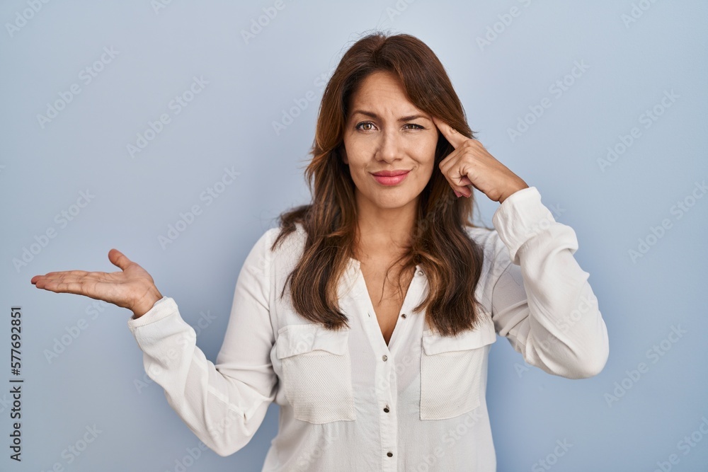 Hispanic woman standing over isolated background confused and annoyed with open palm showing copy space and pointing finger to forehead. think about it. Hispanic woman standing over isolated background confused and annoyed with open palm showing copy space and pointing finger to forehead. think about it.