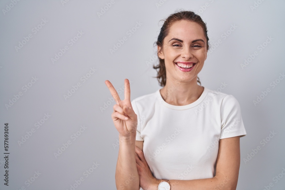 Beautiful brunette woman standing over isolated background smiling with happy face winking at the camera doing victory sign with fingers. number two.