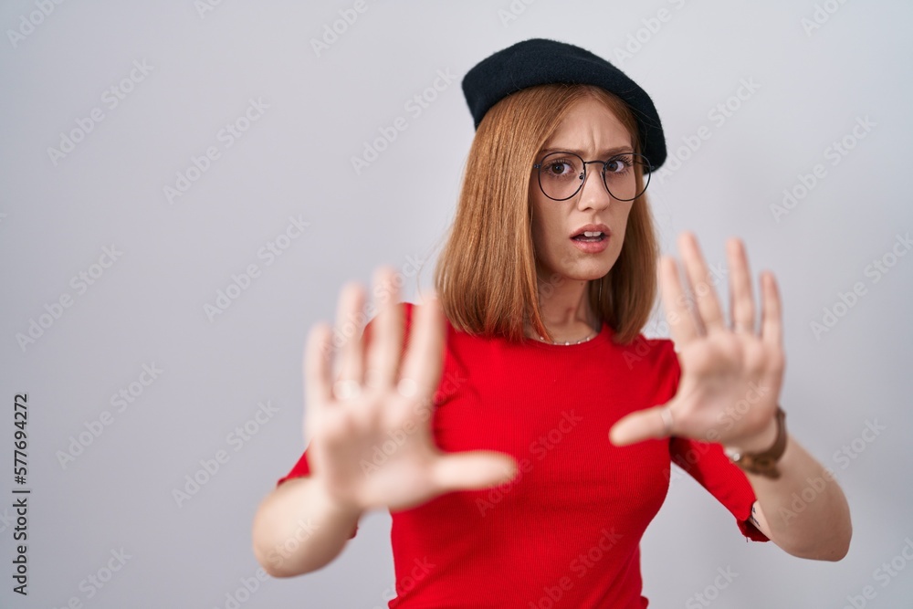 Young redhead woman standing wearing glasses and beret afraid and terrified with fear expression stop gesture with hands, shouting in shock. panic concept. Young redhead woman standing wearing glasses and beret afraid and terrified with fear expression stop gesture with hands, shouting in shock. panic concept.