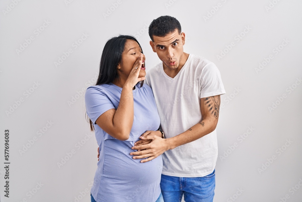 Young hispanic couple expecting a baby standing over background hand on mouth telling secret rumor, whispering malicious talk conversation Young hispanic couple expecting a baby standing over background hand on mouth telling secret rumor, whispering malicious talk conversation