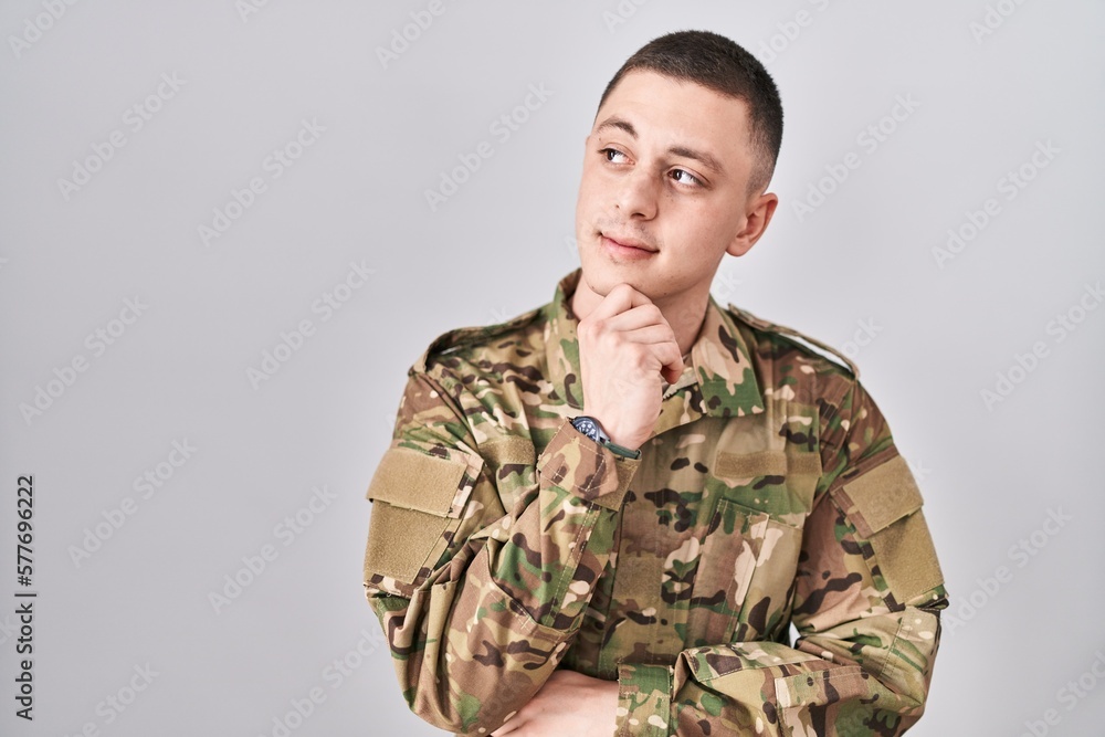 Young man wearing camouflage army uniform with hand on chin thinking about question, pensive expression. smiling with thoughtful face. doubt concept.