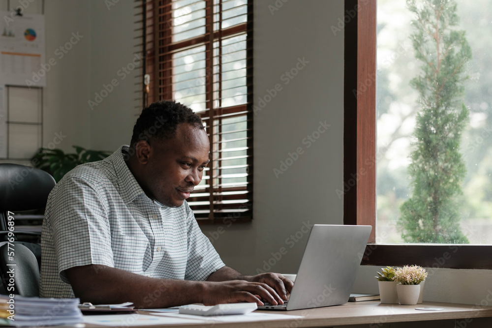Smiling black man using laptop at home in living room. Happy mature businessman send email and working at home. African american freelancer typing on computer with paperworks and documents on table. Smiling black man using laptop at home in living room. Happy mature businessman send email and working at home. African american freelancer typing on computer with paperworks and documents on table.