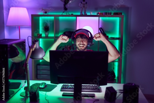 Young hispanic man streamer using computer and headphones with winner expression at gamin room