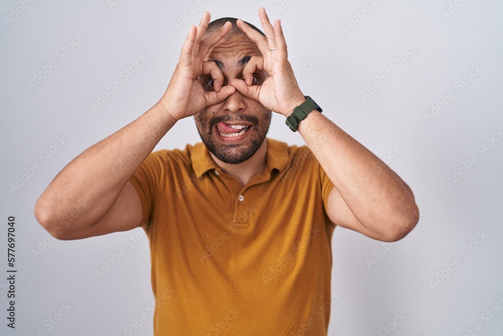 Fototapeta premium Hispanic man with beard standing over white background doing ok gesture like binoculars sticking tongue out, eyes looking through fingers. crazy expression.