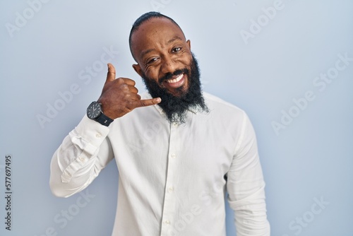 African american man standing over blue background smiling doing phone gesture with hand and fingers like talking on the telephone. communicating concepts.