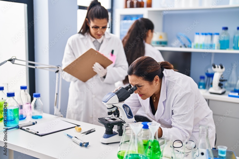 Three woman scientists using microscope write on checklist at laboratory Three woman scientists using microscope write on checklist at laboratory