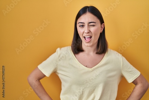 Hispanic girl wearing casual t shirt over yellow background winking looking at the camera with sexy expression, cheerful and happy face.