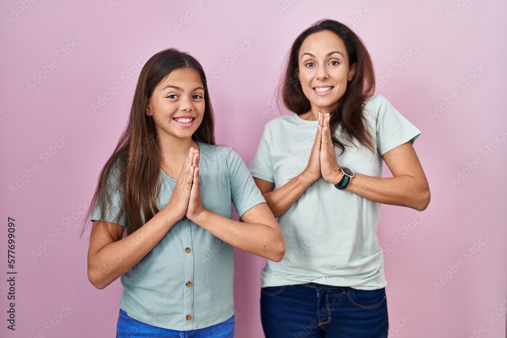 Young mother and daughter standing over pink background praying with hands together asking for forgiveness smiling confident. Young mother and daughter standing over pink background praying with hands together asking for forgiveness smiling confident.