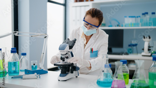 Middle age hispanic woman wearing scientist uniform and medical mask using microscope at laboratory