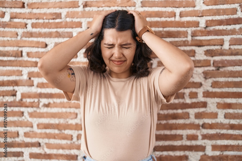 Young hispanic woman standing over bricks wall suffering from headache desperate and stressed because pain and migraine. hands on head. Young hispanic woman standing over bricks wall suffering from headache desperate and stressed because pain and migraine. hands on head.