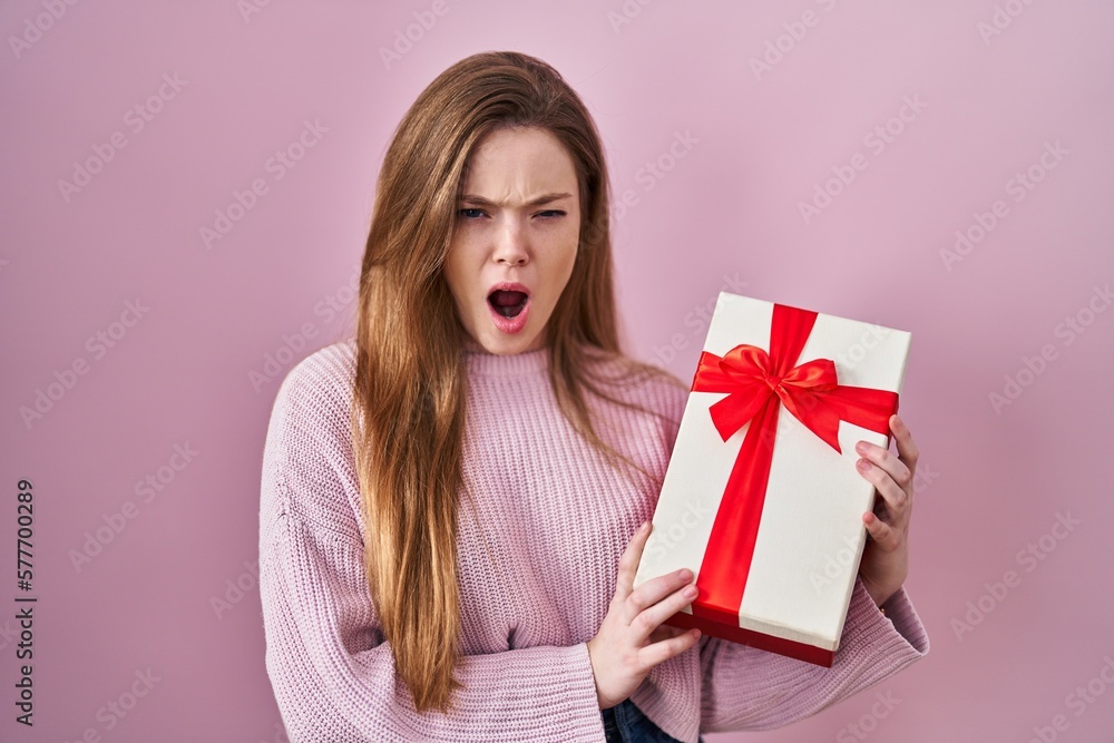 Young caucasian woman holding gift angry and mad screaming frustrated and furious, shouting with anger. rage and aggressive concept. Young caucasian woman holding gift angry and mad screaming frustrated and furious, shouting with anger. rage and aggressive concept.