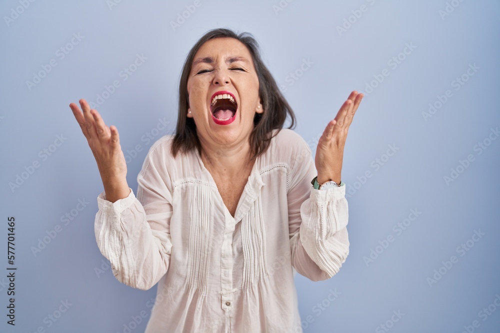 Middle age hispanic woman standing over blue background celebrating mad and crazy for success with arms raised and closed eyes screaming excited. winner concept Middle age hispanic woman standing over blue background celebrating mad and crazy for success with arms raised and closed eyes screaming excited. winner concept