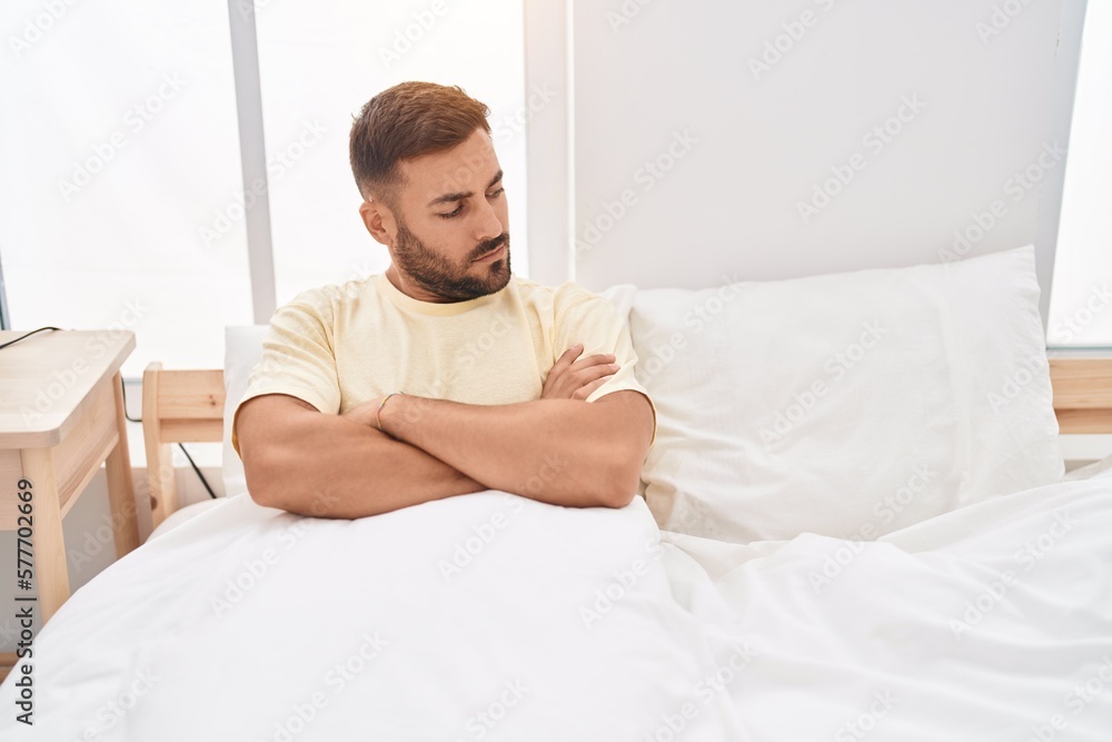 Young hispanic man sitting on bed with unhappy expression and arms crossed gesture at bedroom Young hispanic man sitting on bed with unhappy expression and arms crossed gesture at bedroom
