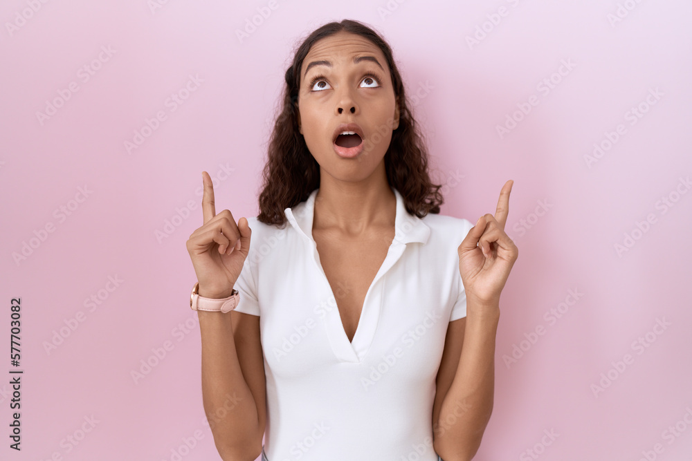 Young hispanic woman wearing casual white t shirt amazed and surprised looking up and pointing with fingers and raised arms. Young hispanic woman wearing casual white t shirt amazed and surprised looking up and pointing with fingers and raised arms.