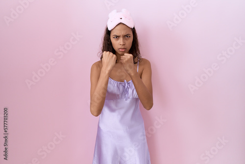 Young hispanic woman wearing sleep mask and nightgown ready to fight with fist defense gesture, angry and upset face, afraid of problem
