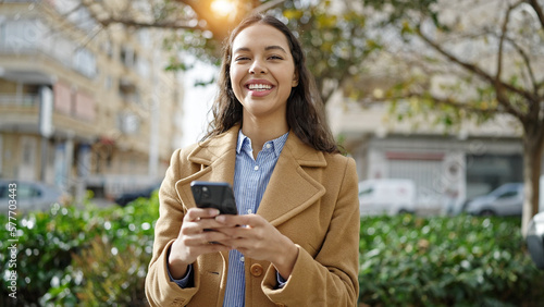 Young beautiful hispanic woman using smartphone smiling at park