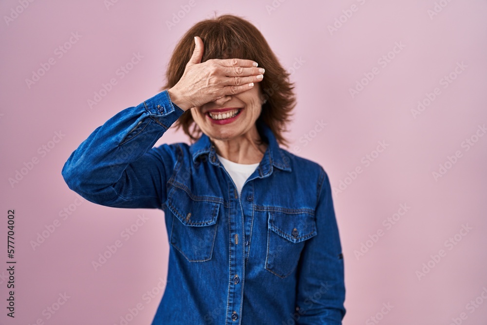 Middle age woman standing over pink background smiling and laughing with hand on face covering eyes for surprise. blind concept. Middle age woman standing over pink background smiling and laughing with hand on face covering eyes for surprise. blind concept.