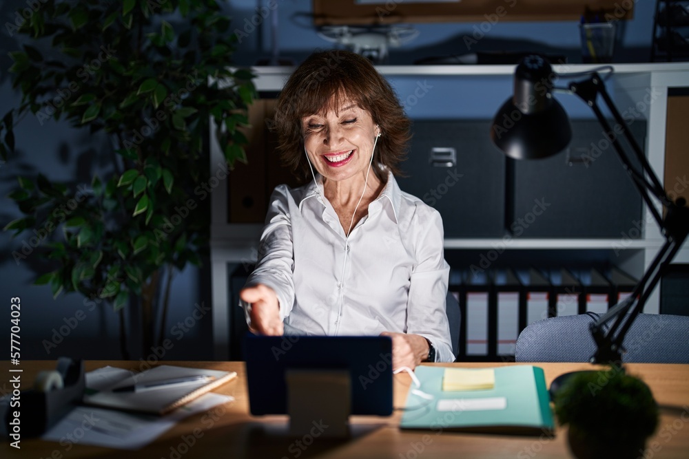 Middle age woman working at the office at night smiling friendly offering handshake as greeting and welcoming. successful business. Middle age woman working at the office at night smiling friendly offering handshake as greeting and welcoming. successful business.
