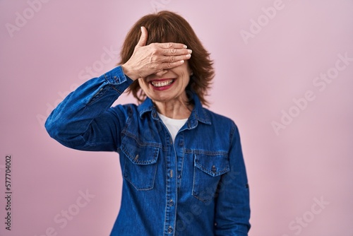 Middle age woman standing over pink background smiling and laughing with hand on face covering eyes for surprise. blind concept. © Krakenimages.com