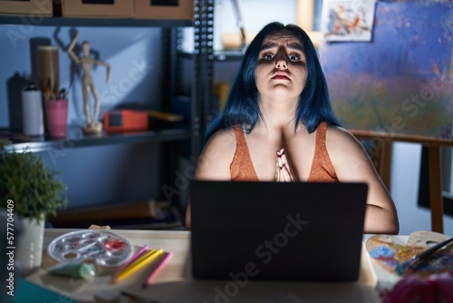 Young modern girl with blue hair sitting at art studio with laptop at night begging and praying with hands together with hope expression on face very emotional and worried. begging.