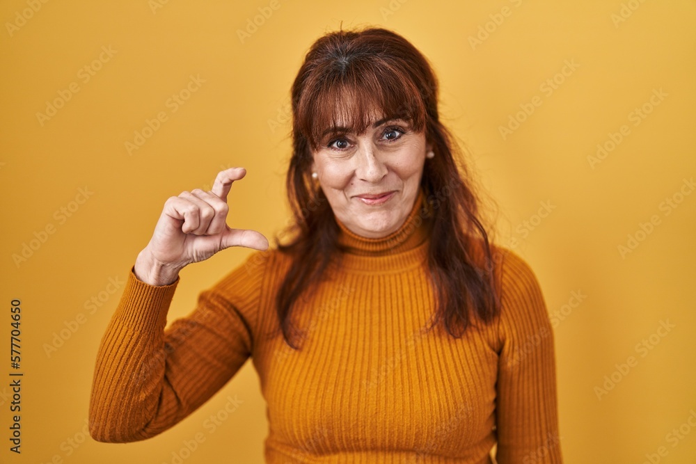 Middle age hispanic woman standing over yellow background smiling and confident gesturing with hand doing small size sign with fingers looking and the camera. measure concept. Middle age hispanic woman standing over yellow background smiling and confident gesturing with hand doing small size sign with fingers looking and the camera. measure concept.