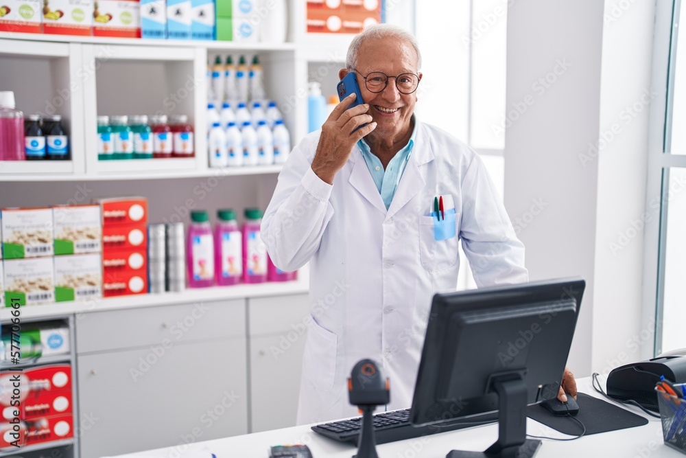 Senior grey-haired man pharmacist talking on smartphone using computer at pharmacy Senior grey-haired man pharmacist talking on smartphone using computer at pharmacy