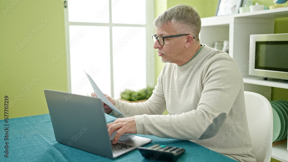 Middle age grey-haired man using laptop reading document at home Middle age grey-haired man using laptop reading document at home