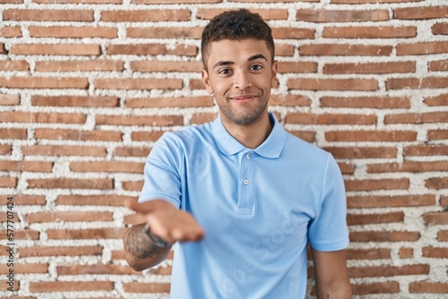 Brazilian young man standing over brick wall smiling cheerful offering palm hand giving assistance and acceptance.