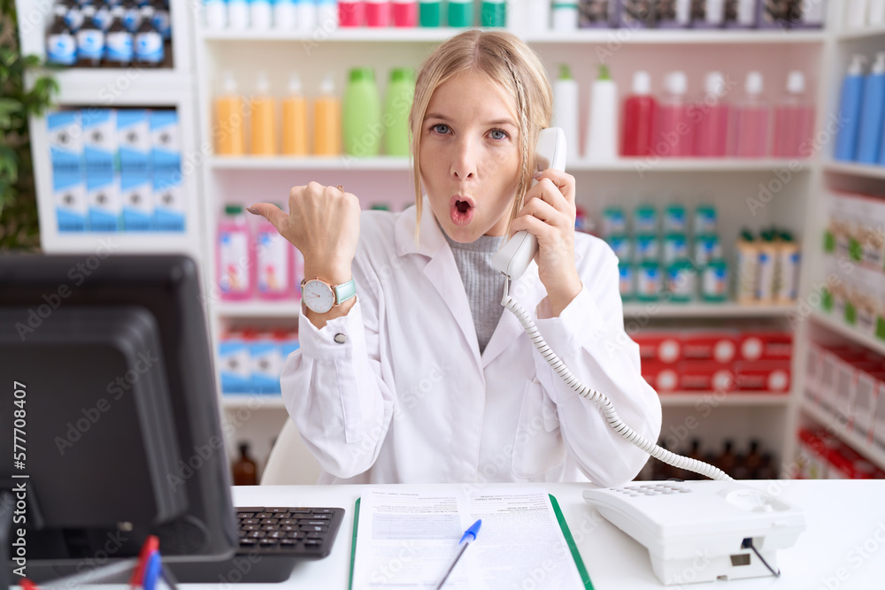 Young caucasian woman working at pharmacy drugstore speaking on the telephone surprised pointing with hand finger to the side, open mouth amazed expression. Young caucasian woman working at pharmacy drugstore speaking on the telephone surprised pointing with hand finger to the side, open mouth amazed expression.