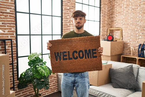 Arab man with beard holding welcome doormat puffing cheeks with funny face. mouth inflated with air, catching air.