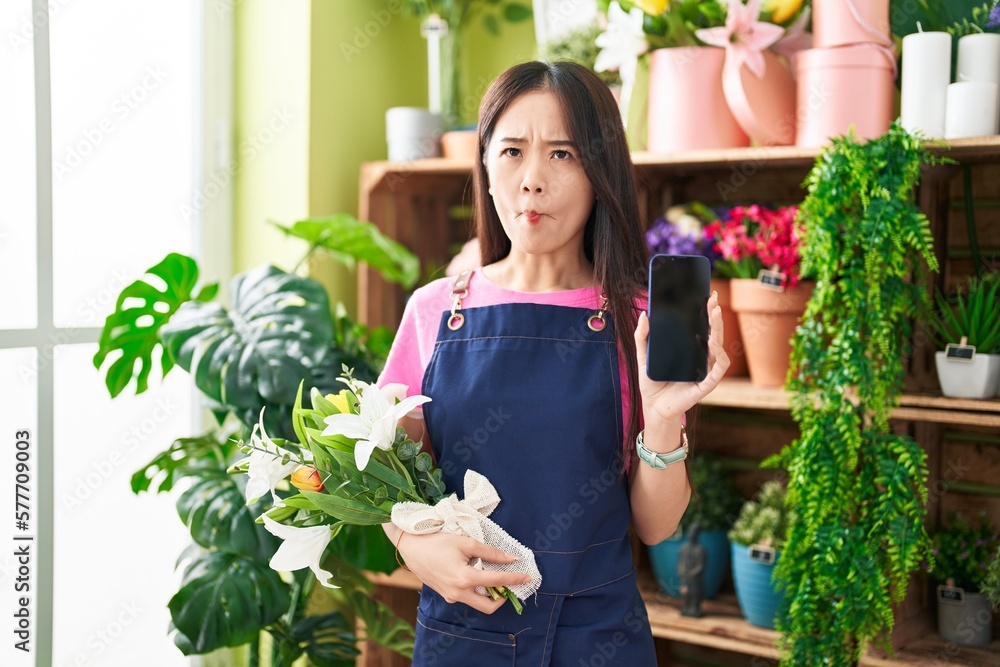 Young chinese woman working at florist shop showing smartphone screen making fish face with mouth and squinting eyes, crazy and comical.